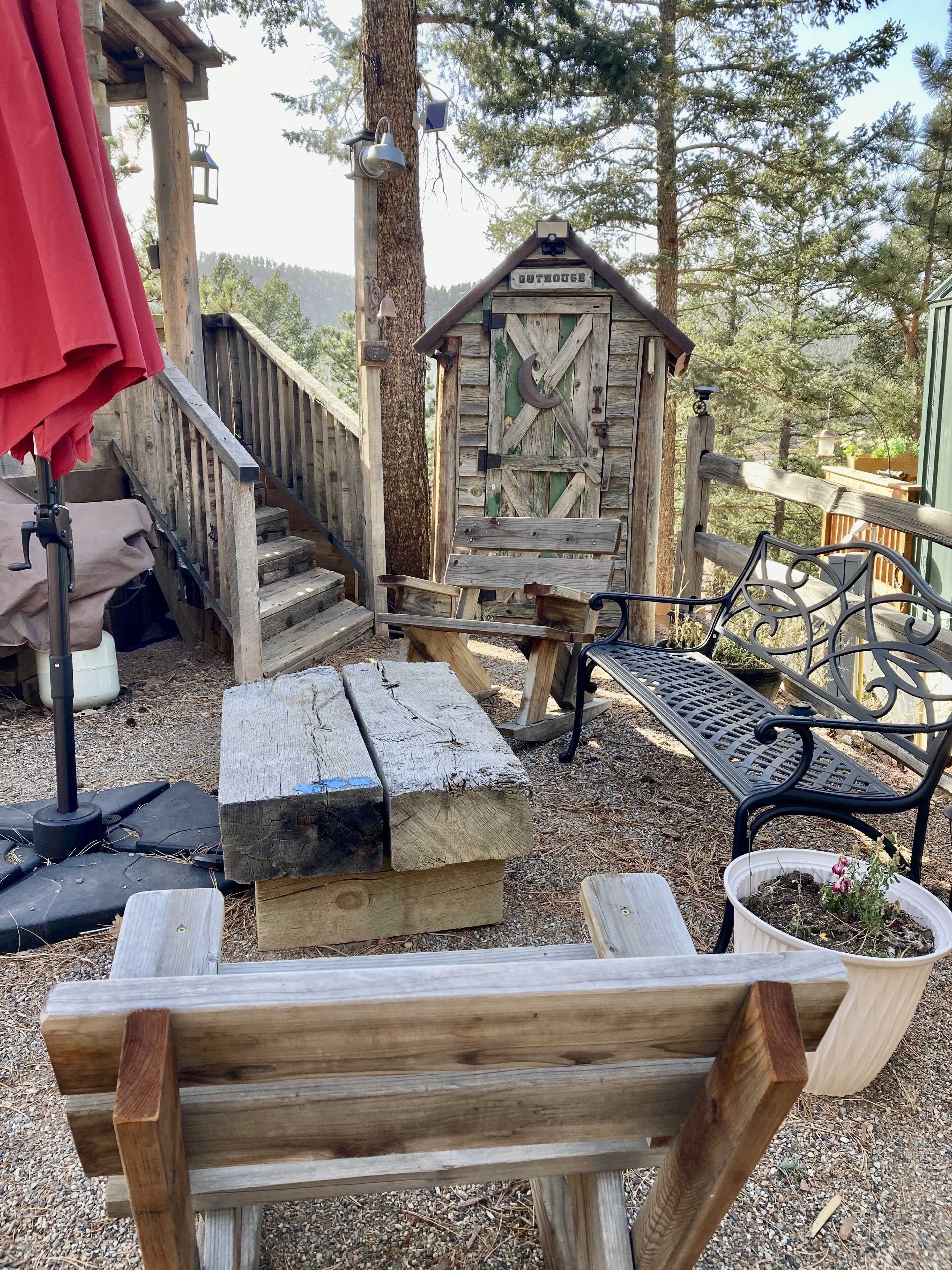 Een rustieke buitenruimte met houten banken en een picknicktafel, omgeven door bomen en een houten constructie met de tekst 'OUTHOUSE'.