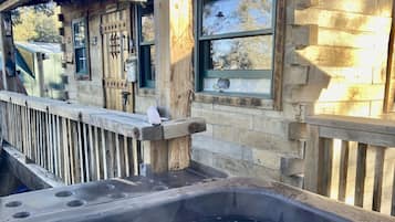 A wooden cabin with a hot tub in front, a wooden deck, and a window with a view of trees.