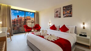 Ein Hotelzimmer mit einem großen Bett, roten Stühlen, einem Schreibtisch und Blick auf die Stadt durch große Fenster.