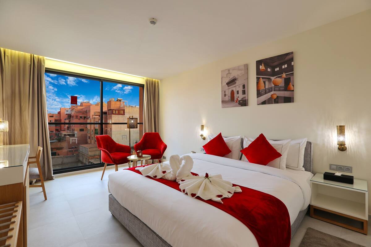 A hotel room with a large bed, red chairs, a desk, and a view of the city through large windows.
