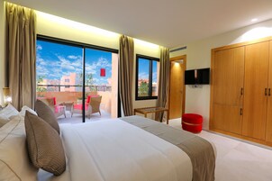 A hotel room with a large bed, a television, a wooden wardrobe, and a balcony with a view of buildings.