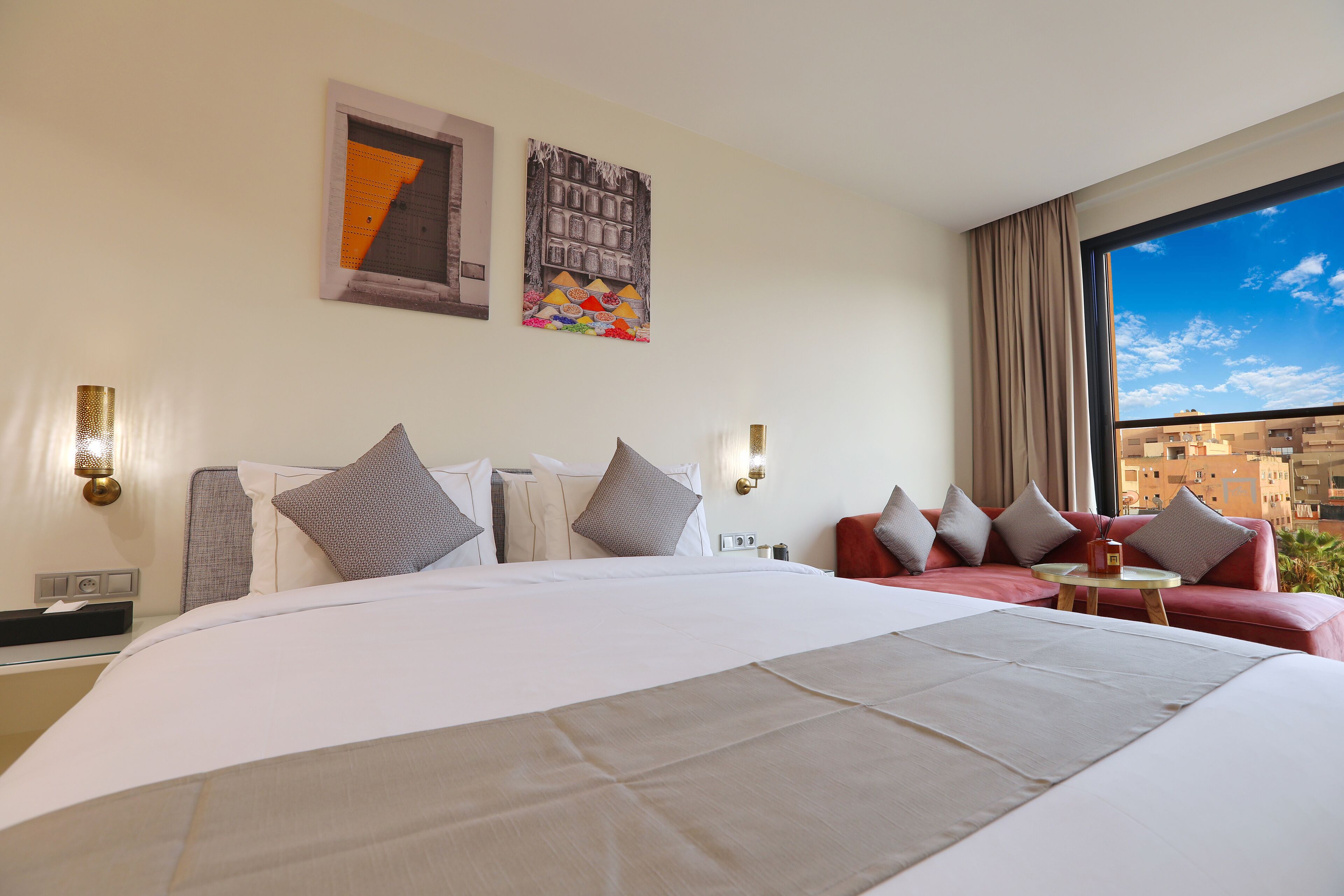Une chambre d’hôtel avec un grand lit, un canapé rouge et une petite table. Il y a deux lampes fixées au mur et une fenêtre donnant sur des immeubles.