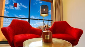 Ein Zimmer mit zwei roten Stühlen, einem Glastisch, einer dekorativen Lampe und Blick auf den Himmel durch große Fenster.