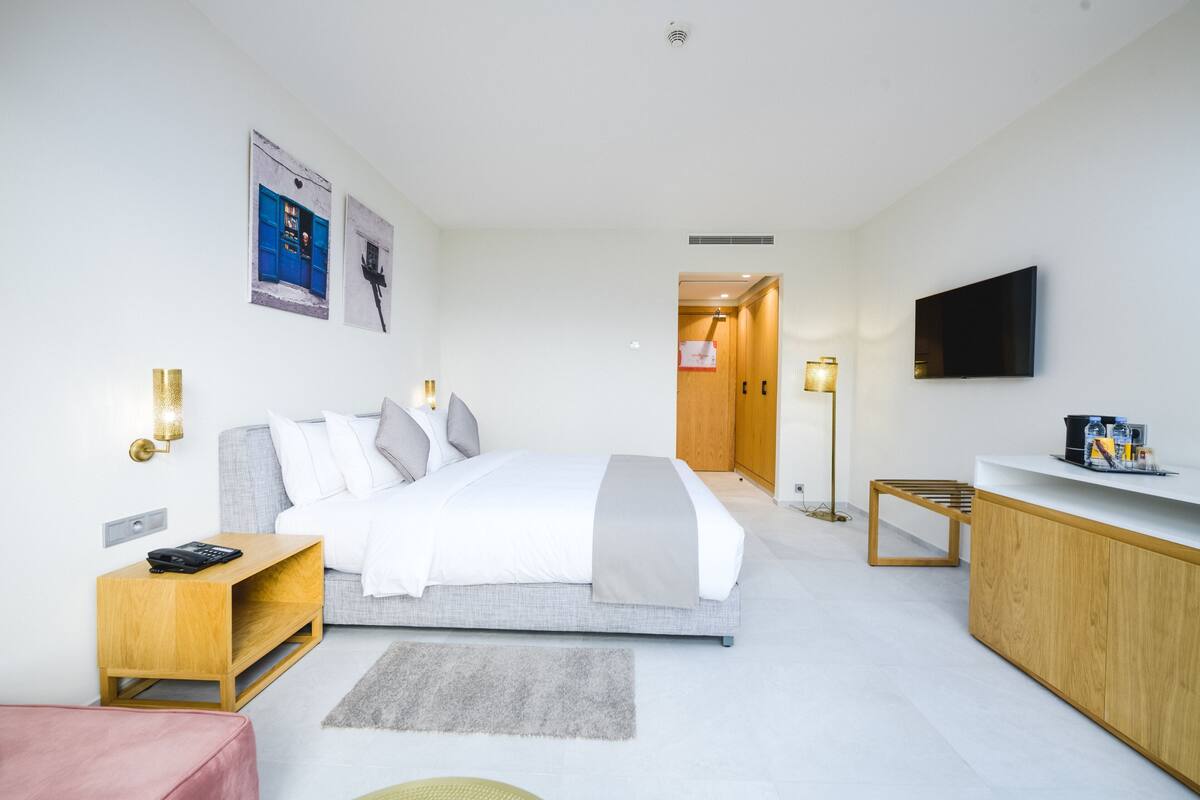 A modern hotel room with a bed, bedside table, TV, and a desk with a chair.