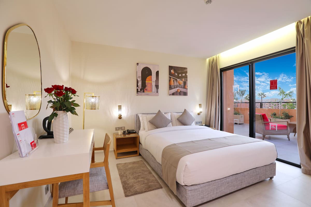 A hotel room with a large bed, a desk with a chair, a mirror, and a view of the outdoors.