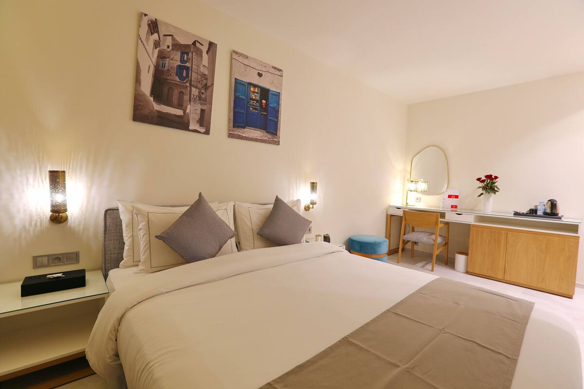 A hotel room with a large bed, a desk, a chair, a small stool, a mirror, and two wall-mounted light fixtures.