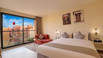 Habitación de hotel con una cama grande, un sillón rojo, una mesita de noche y vistas de la ciudad a través de ventanales de gran altura.