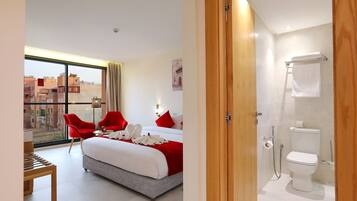 Una habitación de hotel moderna con una cama grande, sillas rojas, una mesa de madera y vistas a la ciudad a través de amplias ventanas.