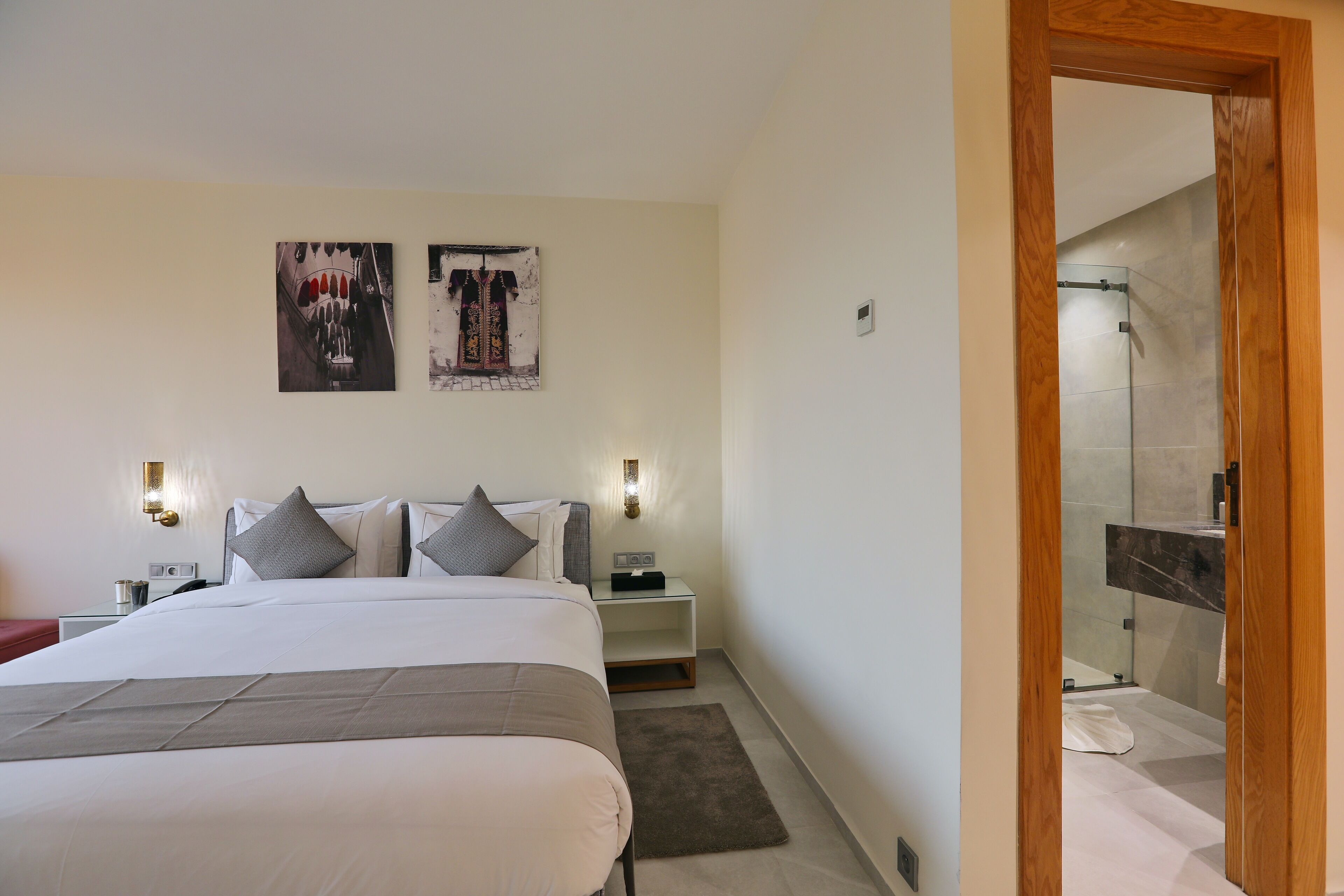 A modern bedroom with a large bed, two bedside tables, and a nightstand. There are two wall-mounted light fixtures and two framed artworks above the bed.