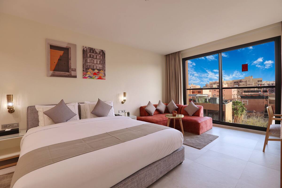 A modern hotel room with a large bed, a red sofa, a small table, and a large window offering a city view.