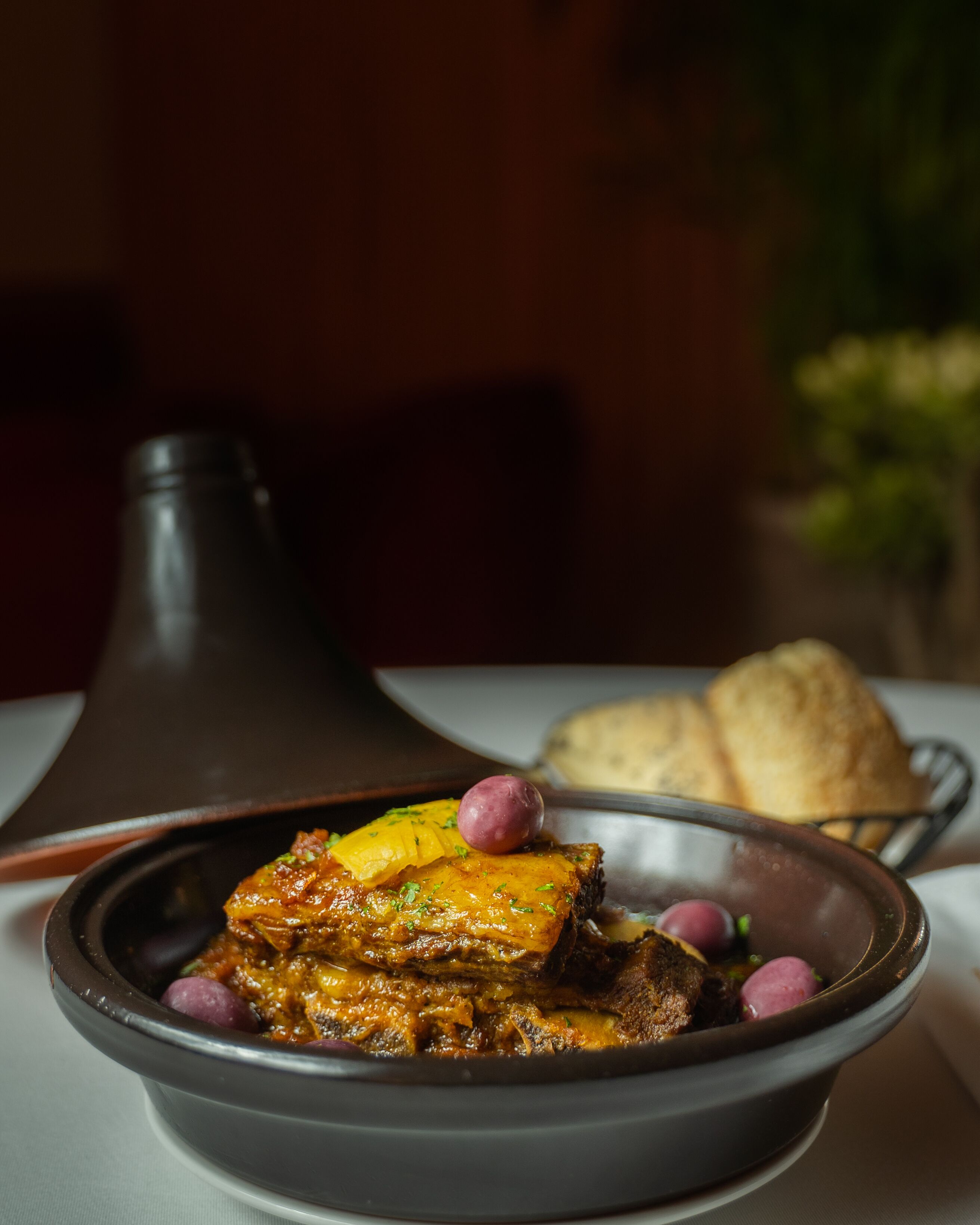 Un tazón de estofado con carne y verduras, acompañado de un recipiente pequeño de salsa y una rebanada de pan sobre una mesa.