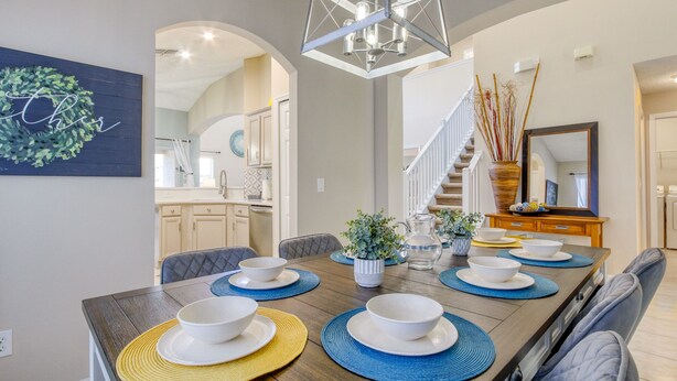 Une salle à manger avec une longue table dressée pour six personnes, un lustre et un escalier menant à un autre niveau.