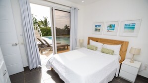 Ein Schlafzimmer mit einem großen Bett, einem Nachttisch, einer Lampe und Blick auf einen Pool und Palmen.