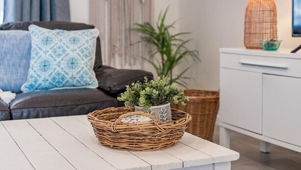 Una cesta de mimbre con una planta sobre una mesa blanca en una sala de estar con un sofá, cojines, una lámpara y un aparador.