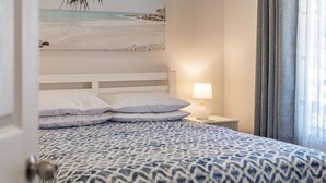Un dormitorio con una colcha estampada, una lámpara de noche y un cuadro con una playa.