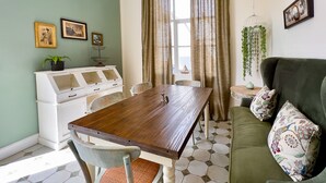A dining area with a wooden table, chairs, and a white cabinet with glass doors. There is a green sofa with floral cushions and a hanging plant. - Brigadoon Boutique Guesthouse l Ondili (Swakopmund)