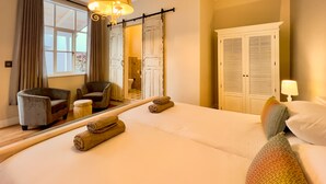 A hotel room with a large bed, two armchairs, a small round table, a mirror, and a wardrobe with louvered doors. - Brigadoon Boutique Guesthouse l Ondili (Swakopmund)