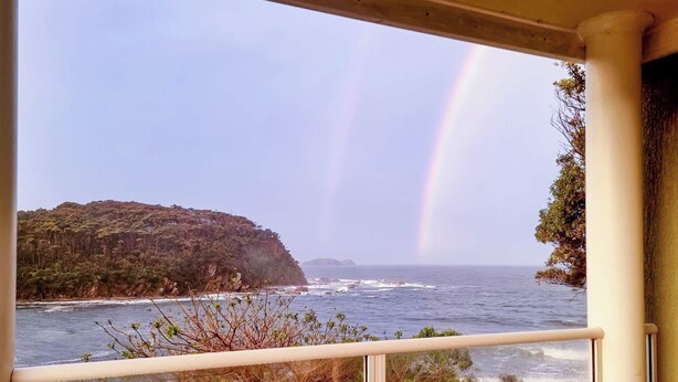 A view from a balcony overlooking a coastal landscape with a rainbow, a rocky cliff, and the ocean.