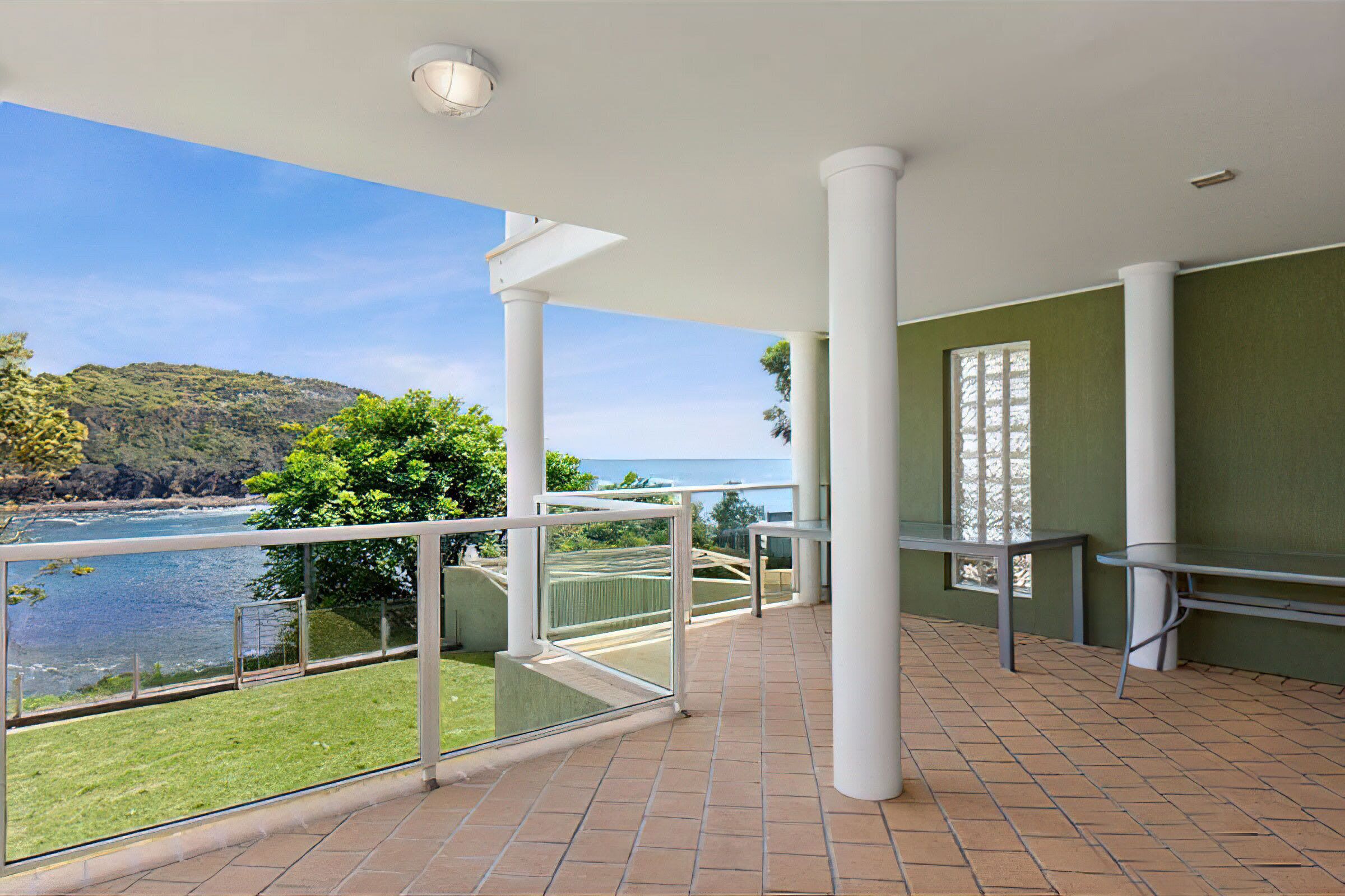 A balcony with a view of the sea and hills, white columns, and a glass railing.