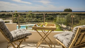 A balcony with two cushioned chairs, a small table with a drink and fruit, and a view of a pool and landscape. - Queen Bee Residence- Queen Apartment (Minia, Kefalonia)