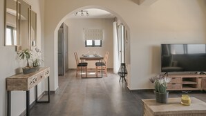 A modern living room with a flat-screen TV, a dining area with a table and chairs, and a console table with decorative items. - Queen Bee Residence- Queen Apartment (Minia, Kefalonia)
