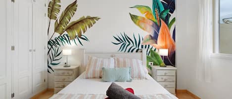 A bedroom with a bed, nightstands, a large tropical mural, and a window.