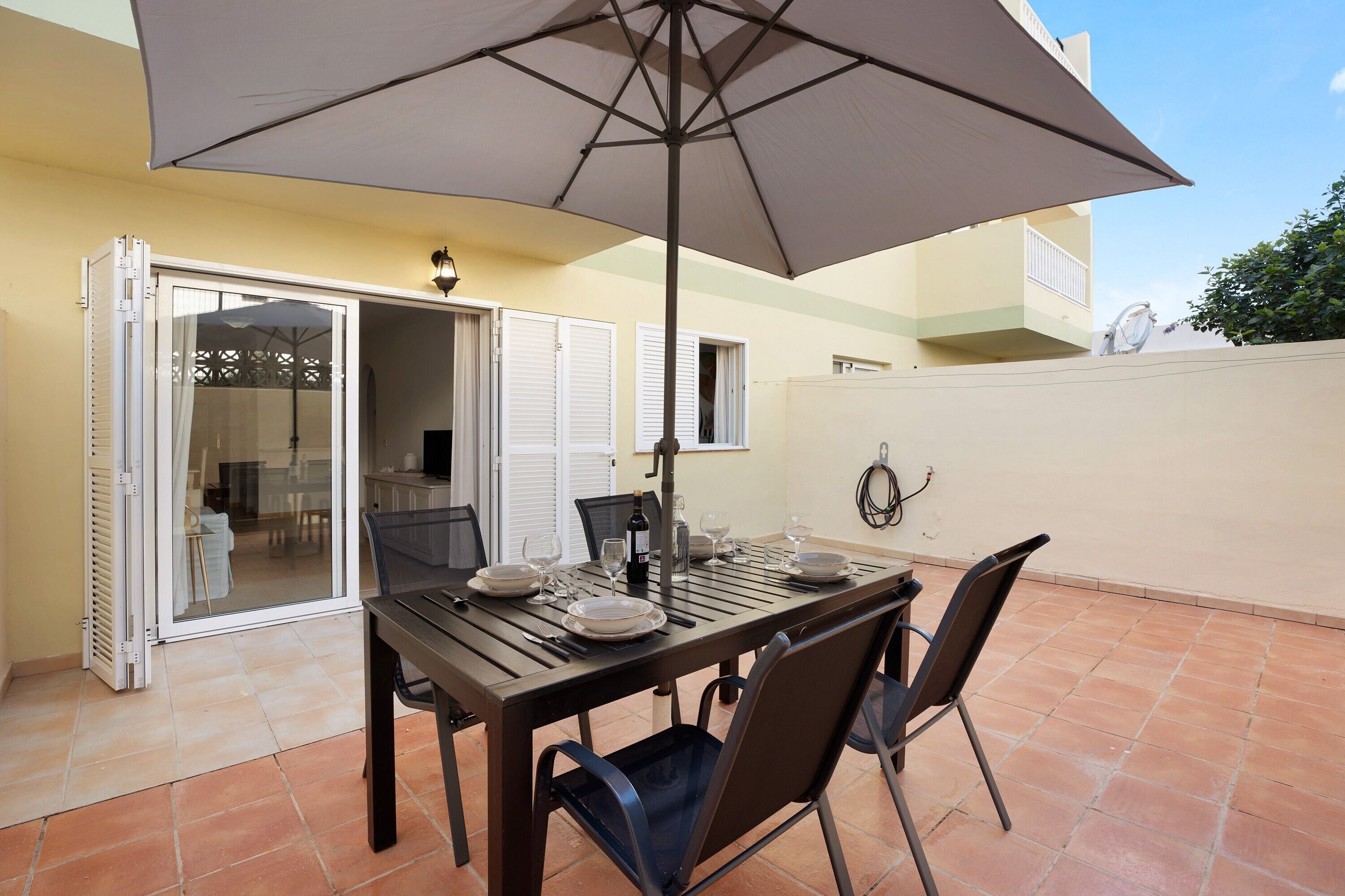 Un'area pranzo all'aperto con tavolo, sedie, ombrellone e una porta scorrevole in vetro che dà su un ambiente interno.