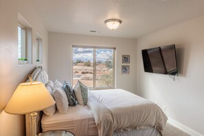 A bedroom with a bed, a bedside lamp, a TV mounted on the wall, and a window with a view of the desert. - Contemporary 4BR w/ 2 Kings - Near 18th Fairway (St. George)