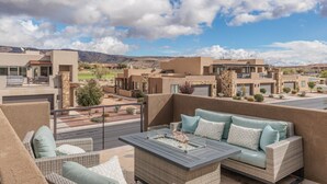 Un balcon doté de meubles en osier et d’une table à plateau de verre, offrant une vue sur un quartier résidentiel avec des maisons modernes et un ciel dégagé.