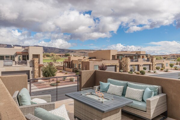 Un balcón con mobiliario de mimbre y una mesa de vidrio, con vistas a una zona residencial con casas modernas y un cielo despejado.