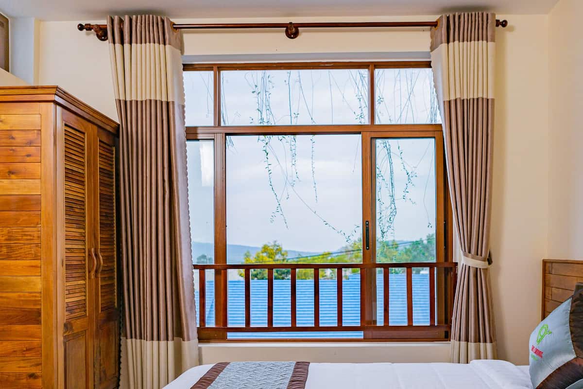 A bedroom with a large window, wooden wardrobe, and a bed with pillows.