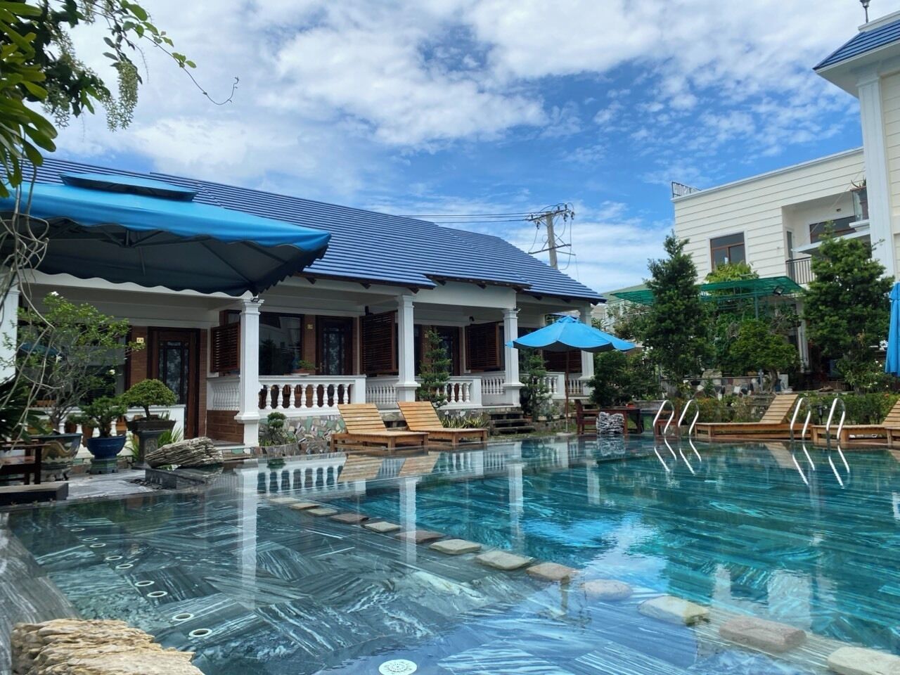 A swimming pool with lounge chairs and umbrellas, surrounded by a building with a covered porch and a white exterior.