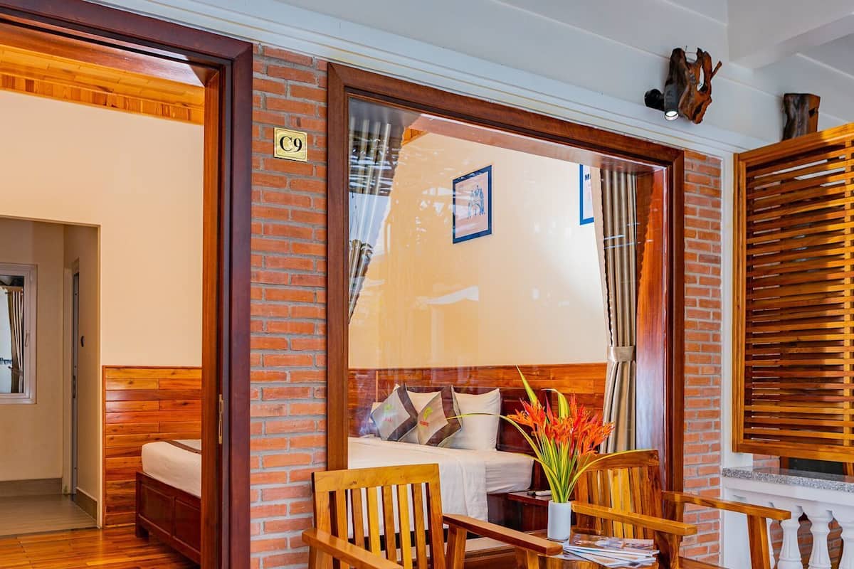 A room with a wooden bed, a wooden chair, a table with a vase, and a brick wall.