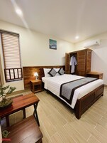 Quarto de hotel com cama de madeira, mesa de cabeceira, escrivaninha, planta em vaso e guarda-roupa.