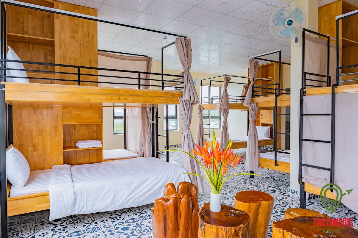 A dormitory room with bunk beds, wooden shelves, a fan, and a decorative vase.