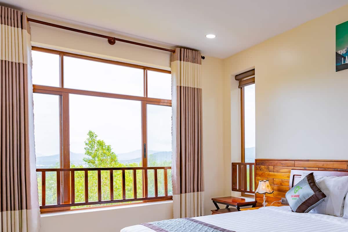 A hotel room with a large window offering a view of greenery and mountains, a wooden headboard, and a painting of a mountain on the wall.