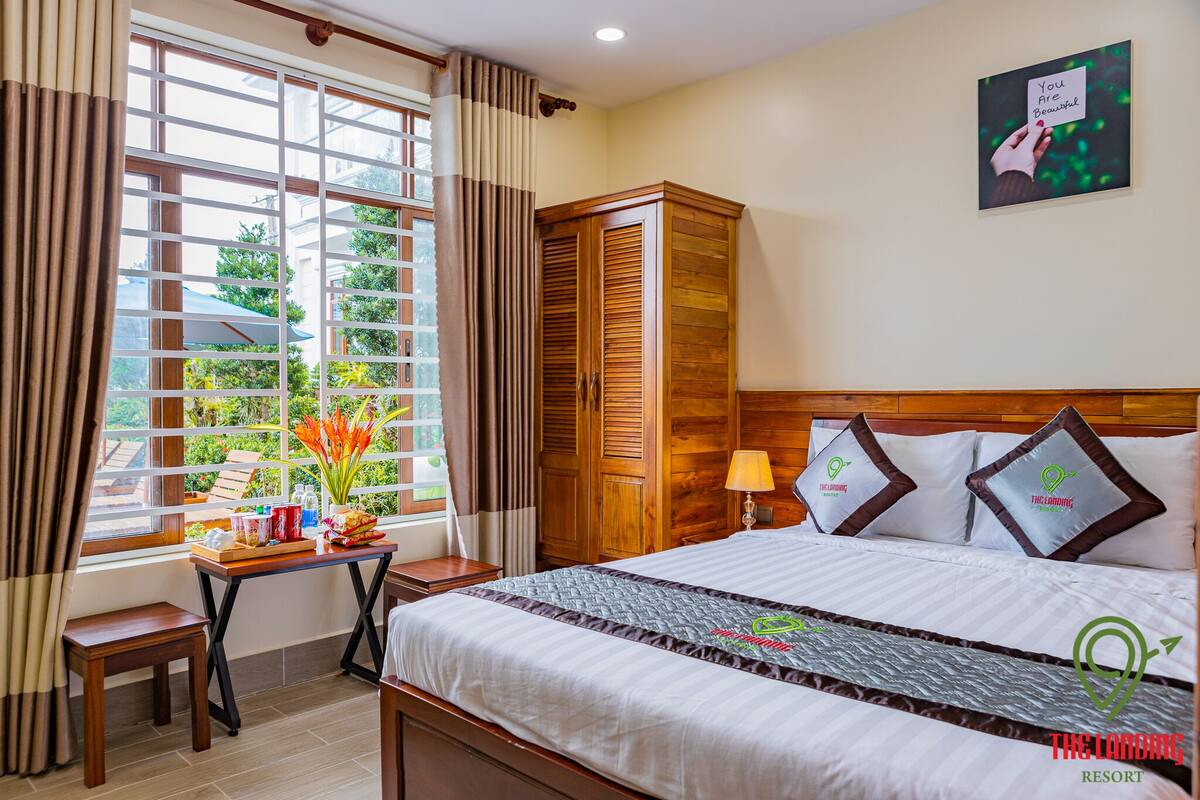 A hotel room with a large window, a wooden wardrobe, a bed with white bedding, and a wall-mounted picture.