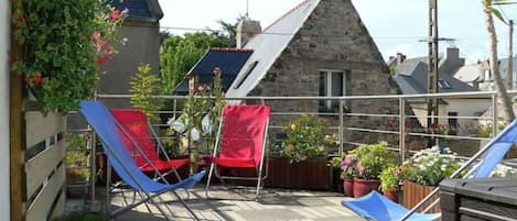 Une terrasse en bois avec deux chaises bleues et une chaise rouge, entourée de plantes en pot et d’un bâtiment en pierre en arrière-plan.