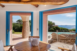 A room with a wooden table, a stone pillar, and a view of a swimming pool and landscape. - Villa Corallina - Porto Cervo (Porto Cervo)