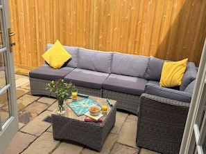 A grey sectional sofa with yellow cushions, a wicker coffee table with books and a bowl of snacks, and a vase with flowers. - 11 Crabbe Street, Aldeburgh (Aldeburgh)