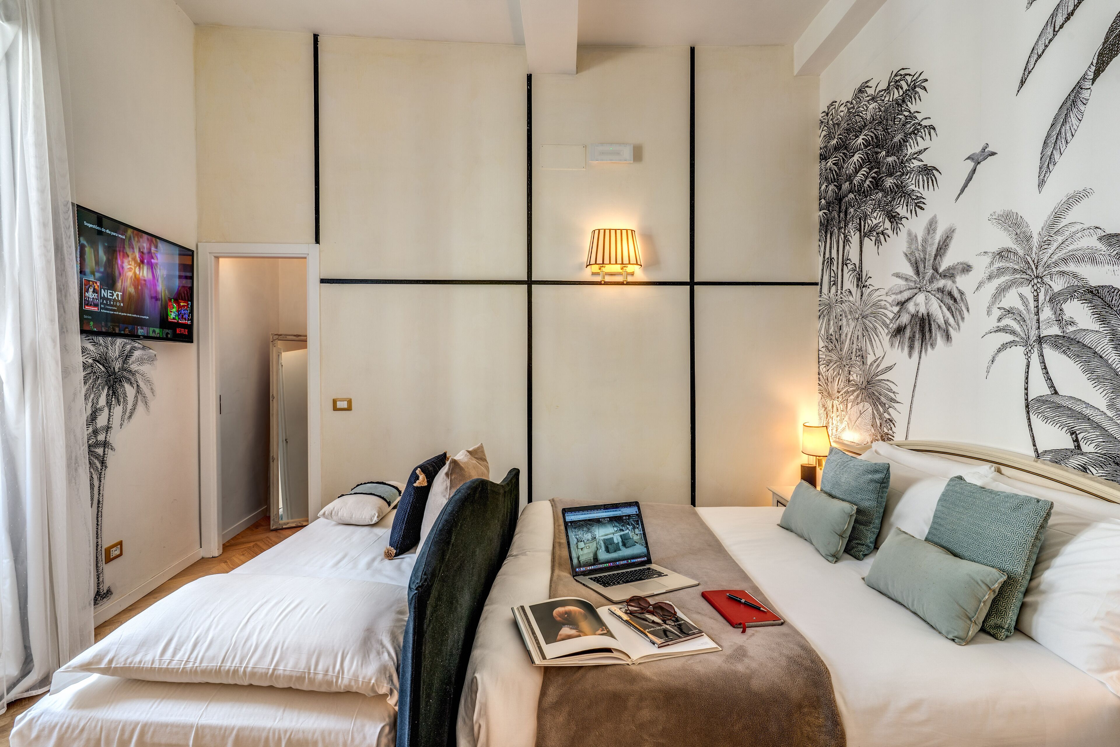 Une chambre d’hôtel avec deux lits, une télévision, un ordinateur portable et un mur orné d’une fresque aux motifs tropicaux.