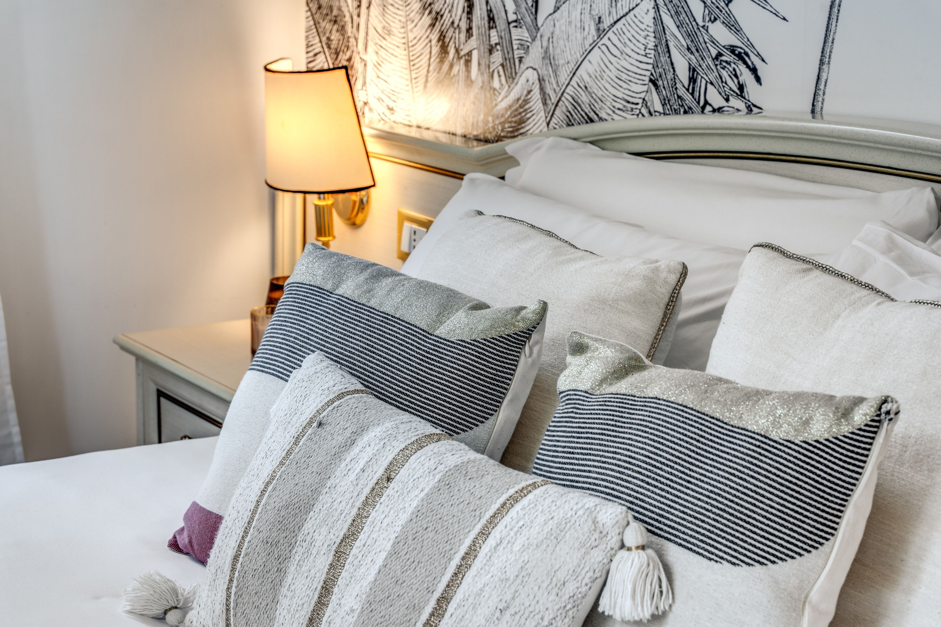 A neatly made bed with striped and plain pillows, a bedside lamp, and a wall with a leaf pattern.