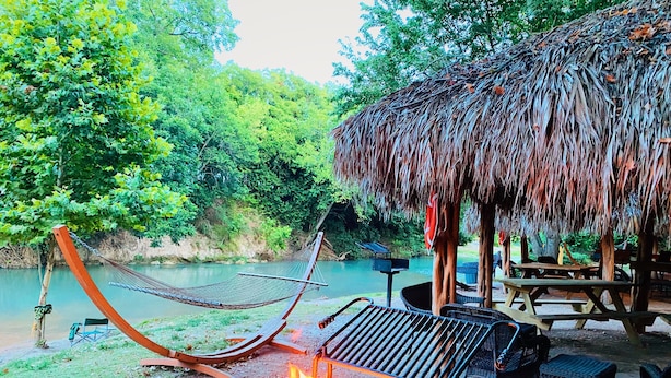 A hammock, a fire pit, and a thatched-roof structure by a river. - Son's River Ranch Glamping Cabin 14 (Kingsbury)