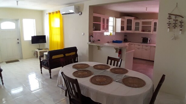 Uma área de refeições com uma mesa redonda para quatro pessoas, uma televisão em um suporte e uma cozinha visível por uma porta aberta.
