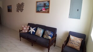Uma sala de estar com um sofá de cor escura, uma cadeira de madeira e uma mesa de jantar com toalha branca. Há várias decorações na parede, incluindo um quadro com uma cena de praia e uma obra de arte com padrão geométrico.