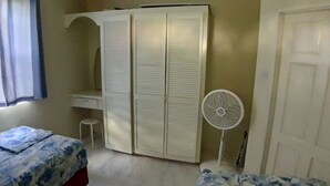 Quarto com cama, guarda-roupa, um pequeno banquinho e ventoinha.