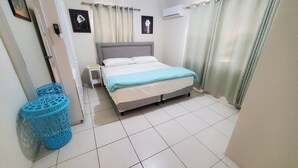 A bedroom with a bed, a nightstand, a wall-mounted air conditioner, and a window with curtains. - Oistins 2 Bed Apartment with AC Near Miami Beach & Oistins. (Christ Church)