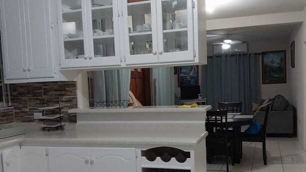 A kitchen with white cabinets, a tiled floor, and a dining area with a table and chairs. - Oistins 2 Bed Apartment with AC Near Miami Beach & Oistins. (Christ Church)