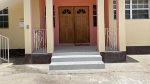 A building entrance with a double wooden door, a staircase leading up, and a metal railing on the left. - Oistins 2 Bed Apartment with AC Near Miami Beach & Oistins. (Christ Church)