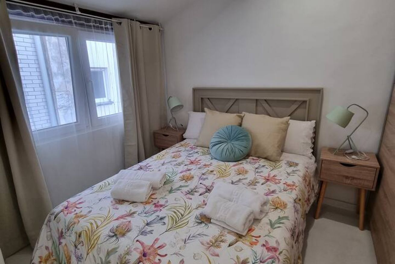 Una cama bien tendida con ropa de cama floral, un cabecero, dos mesitas de noche con lámparas y una ventana con cortinas.